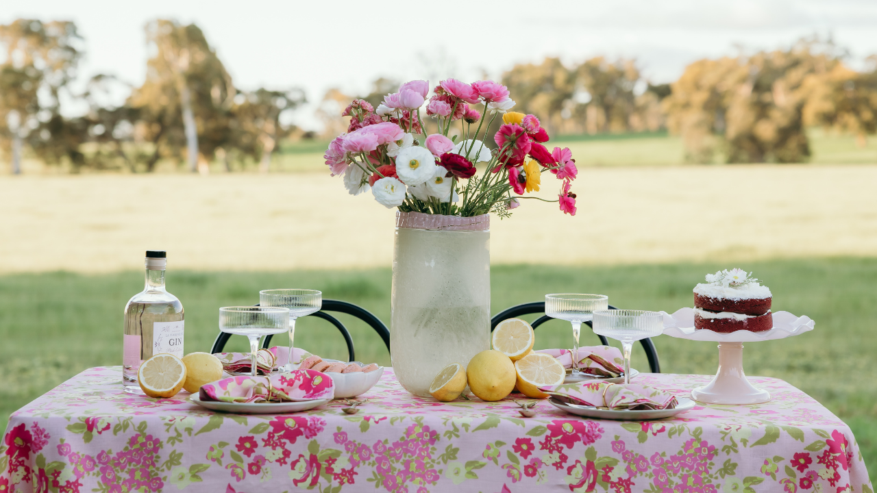 Outdoor setting with a table set for a meal, featuring a cake, flowers, and drinks.